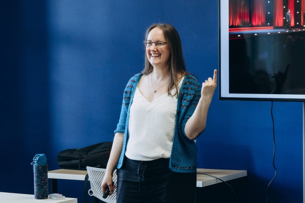 Lizzie is standing and presenting. She's grinning and her left hand is pointing upwards.