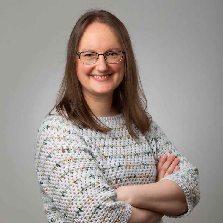Head shot of Lizzie, with her arms folded, smiling at the camera. She's got long brown hair, glasses, and is wearing a pale grey stripey crochet jumper.