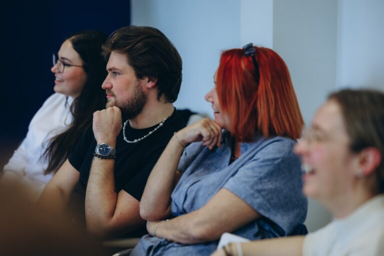 Four people sat in a line at a table, smiling towards a person off camera. Two people have their hands on their chins as if thinking.